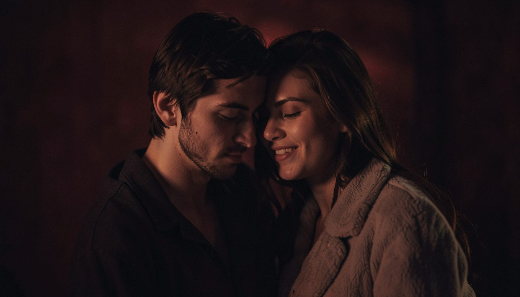 Two figures close together in dim warm light, deep crimson and charcoal tones, intimate and romantic mood
