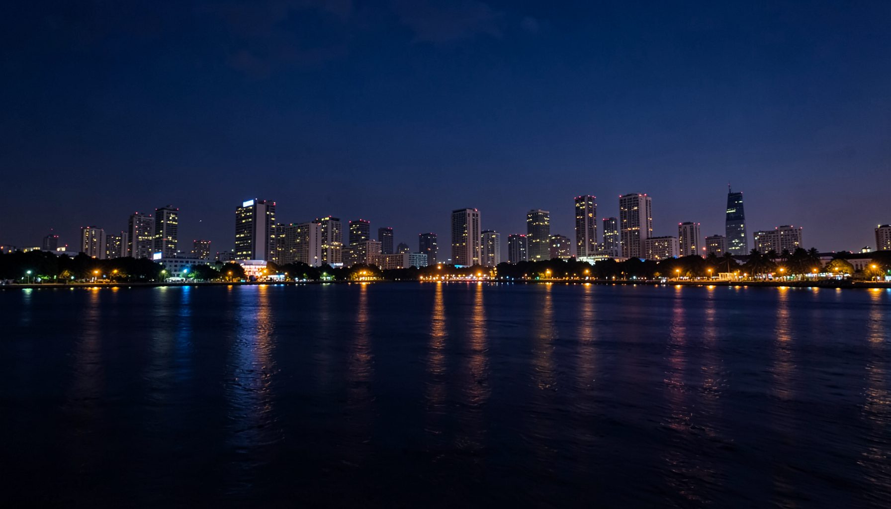 Warm evening city lights reflecting off the waters of Navi Mumbai, deep navy blues and soft amber glows blending across the skyline